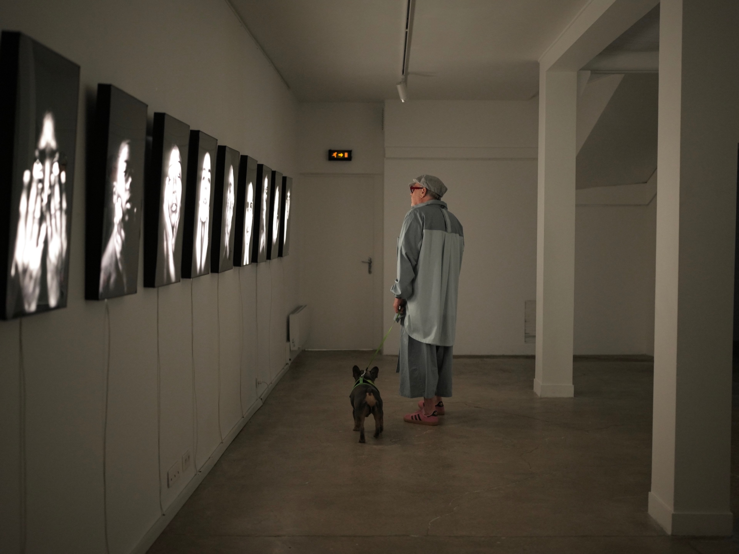 A visitor in pale coveralls with a small dog on a leash, standing in the corridor between the two walls of lightboxes.