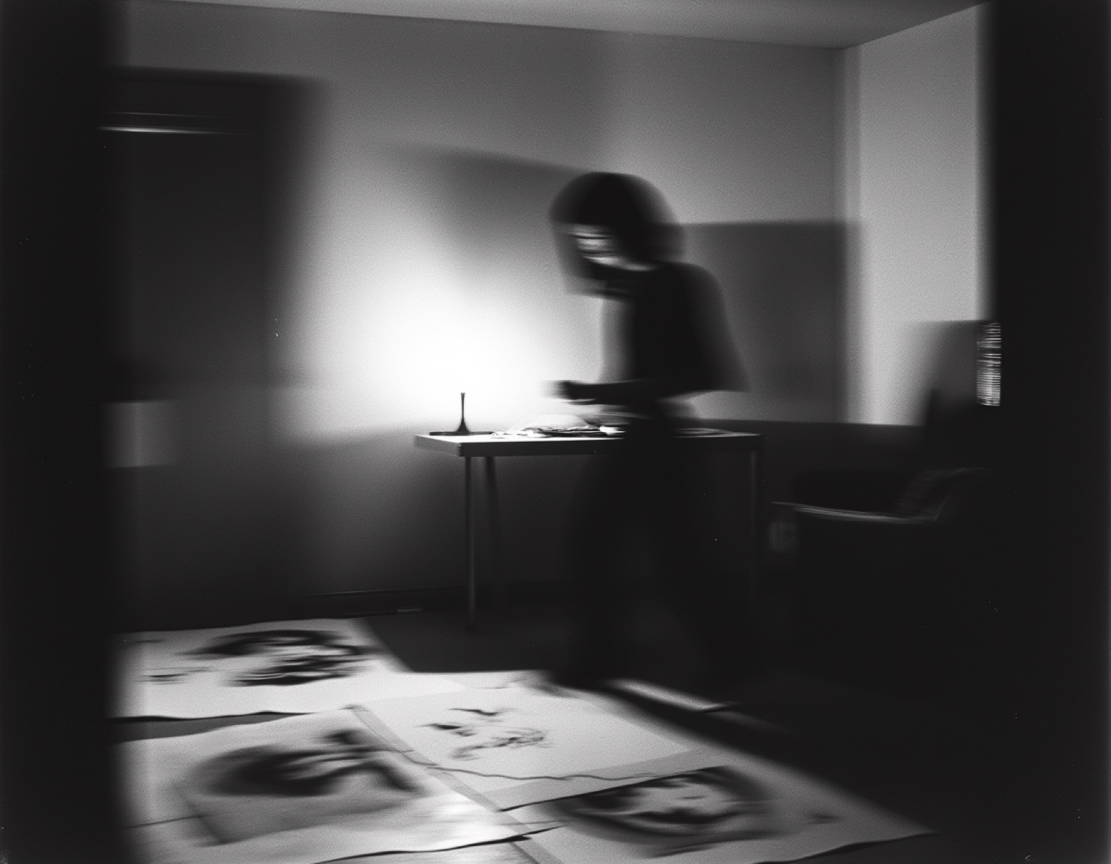 A single figure bent over a lit table in a darkened studio, portraits scattered on the floor.