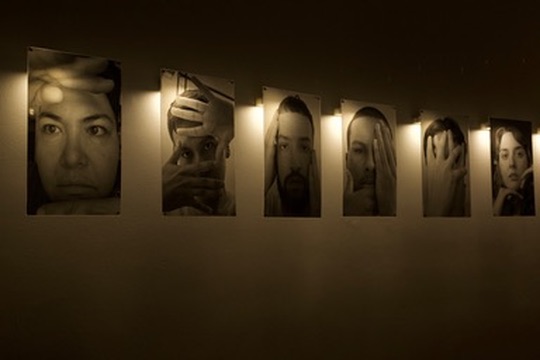 One wall of the corridor: six lit portraits of human subjects, each face partially obscured by a hand. The SPECIMENS.