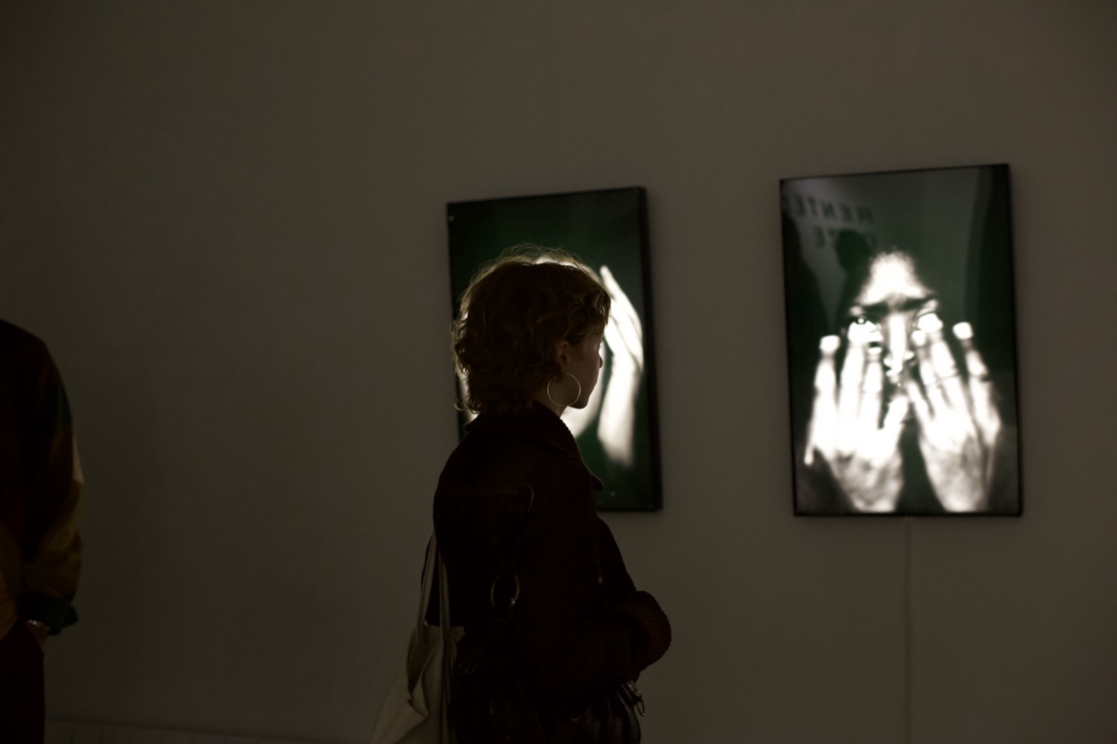 A visitor in silhouette before two green-tinted lightbox portraits — a face framed by hands.